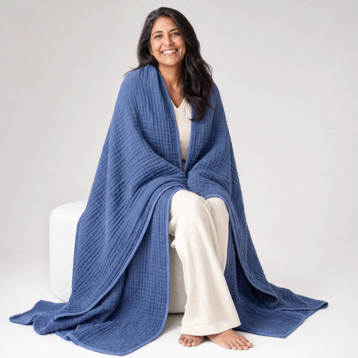 A woman seated beside a bright window with a Dune muslin cotton blanket draped lightly over a wooden chair