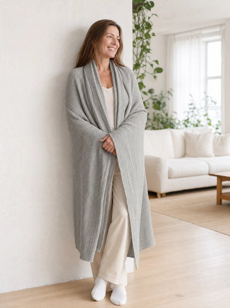 A woman standing wrapped in a grey muslin cotton blanket like a shawl, against a white plaster wall on pale oak flooring