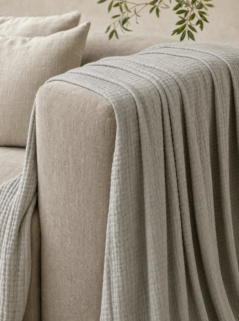 A grey muslin cotton blanket draped over the arm and seat of a linen sofa, falling in deep folds