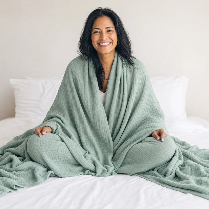 A woman sitting cross-legged on a bed with a sage green muslin cotton blanket wrapped around her shoulders