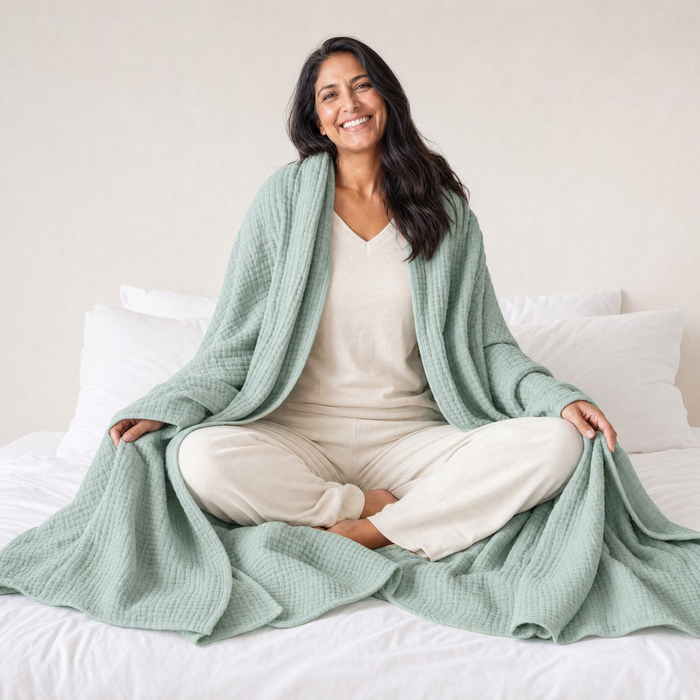Una mujer sentada con las piernas cruzadas en una cama, con una manta de muselina de algodón color verde salvia envuelta alrededor de los hombros