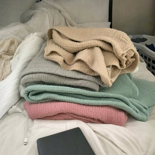 Customer photo: four folded Muslin Blankets in Dune, Grey, Green and Pink on a bed