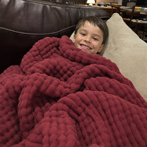 Customer photo: young boy wrapped in the Muslin Blanket in Wine Red on a leather sofa