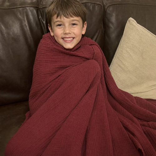 Customer photo: young boy wrapped in the Muslin Blanket in Wine Red on a leather sofa