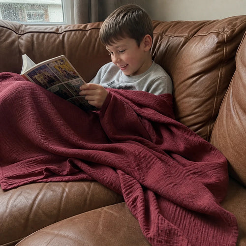 Customer photo: young boy wrapped in the Muslin Blanket in Wine Red on a leather sofa